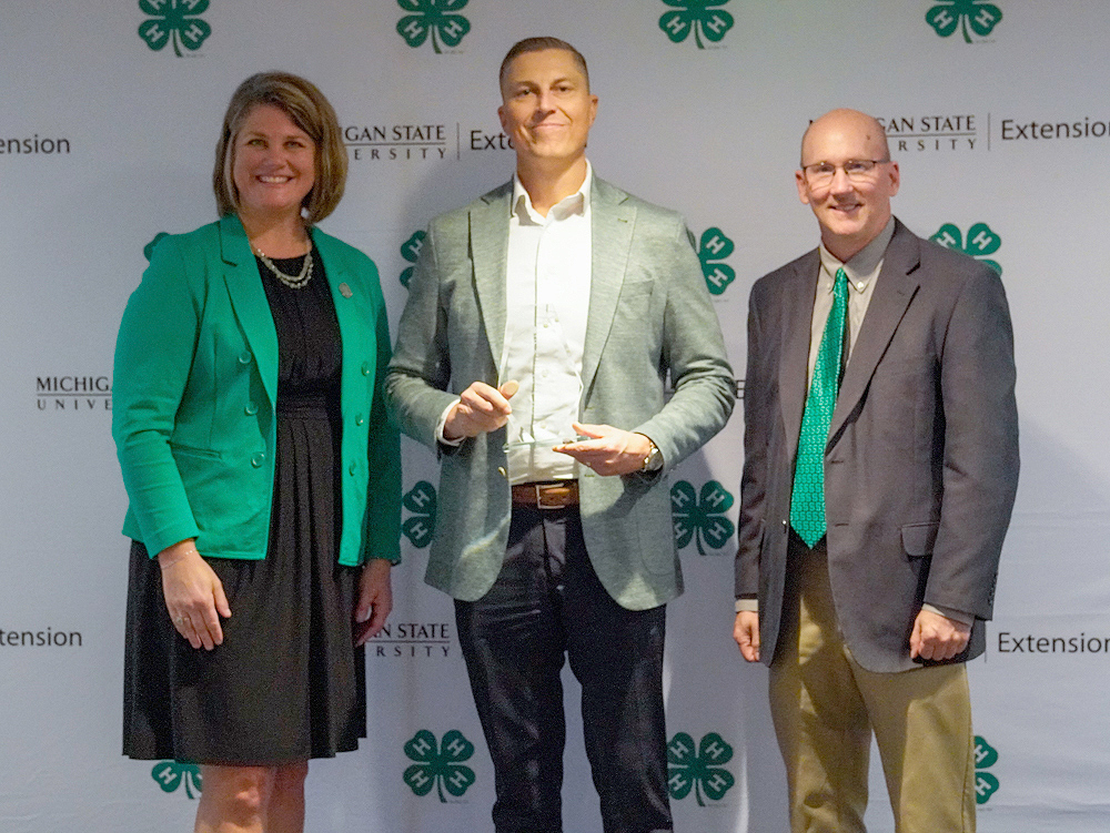 Three individuals at a backdrop. The center person holds an award.
